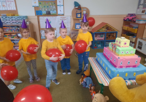 Dzieci w żółtych koszulkach i balonami w ręku stoją przy dużym torcie.