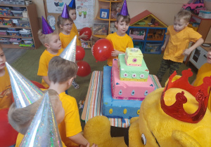 Dzieci w żółtych koszulkach i balonami w ręku stoją przy dużym torcie.
