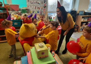 Dzieci w żółtych koszulkach i balonami w ręku wręczają Kubusiowi Puchatkowi baryłkę miodu.