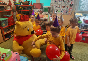 Dzieci w żółtych koszulkach i balonami w ręku wręczają Kubusiowi Puchatkowi baryłkę miodu.