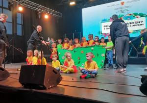 Dzieci występują na dużej scenie w czerwono żółtych strojach.