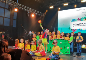 Dzieci występują na dużej scenie w czerwono żółtych strojach.