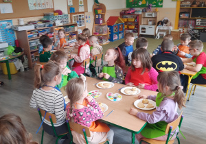 Dzieci w kolorowych fartuchach siedzą przy stolikach i dekorują wielkanocne smakołyki.