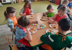 Dzieci w kolorowych fartuchach siedzą przy stolikach i dekorują wielkanocne smakołyki.