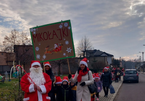 Dzieci z Mikołajem na przodzie, w czapkach mikołajkowych biorą udział w przemarszu ulicami Ozorkowa.