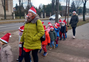 Dzieci z Mikołajem na przodzie, w czapkach mikołajkowych biorą udział w przemarszu ulicami Ozorkowa.