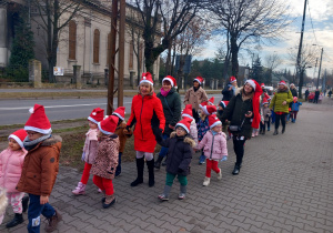 Dzieci z Mikołajem na przodzie, w czapkach mikołajkowych biorą udział w przemarszu ulicami Ozorkowa.