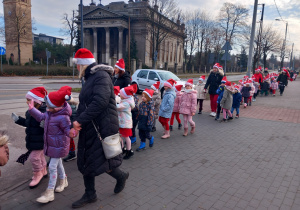 Dzieci z Mikołajem na przodzie, w czapkach mikołajkowych biorą udział w przemarszu ulicami Ozorkowa.