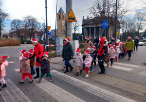 Dzieci z Mikołajem na przodzie, w czapkach mikołajkowych biorą udział w przemarszu ulicami Ozorkowa.