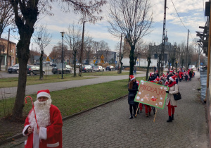 Dzieci z Mikołajem na przodzie, w czapkach mikołajkowych biorą udział w przemarszu ulicami Ozorkowa.