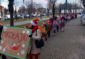 Dzieci z Mikołajem na przodzie, w czapkach mikołajkowych biorą udział w przemarszu ulicami Ozorkowa.