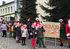 Dzieci z Mikołajem na przodzie, w czapkach mikołajkowych biorą udział w przemarszu ulicami Ozorkowa.