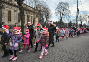 Dzieci z Mikołajem na przodzie, w czapkach mikołajkowych biorą udział w przemarszu ulicami Ozorkowa.