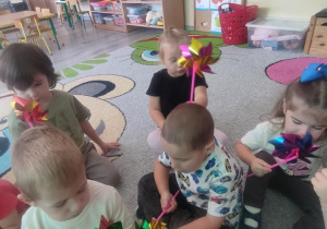 Dzieci na dywanie dmuchają w wiatraczki.