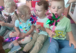 Dzieci na dywanie dmuchają w wiatraczki.