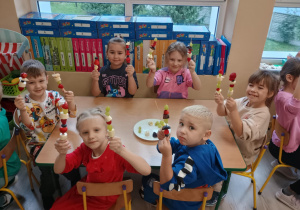 Dzieci przy stoliku siedzą z szaszłykami w rączkach.