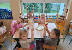Dzieci przy stoliku siedzą z szaszłykami w rączkach.