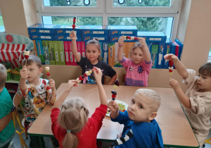 Dzieci przy stoliku siedzą z szaszłykami w rączkach.