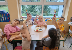 Dzieci przy stoliku siedzą z szaszłykami w rączkach.