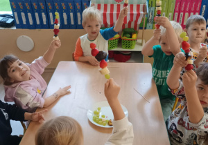 Dzieci przy stoliku siedzą z szaszłykami w rączkach.