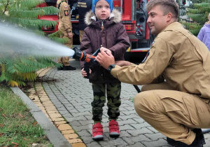 Dziecko wraz z strażakiem leje wodę w prądownicy.