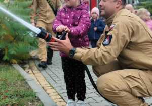 Dziecko wraz z strażakiem leje wodę w prądownicy.
