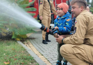 Dziecko wraz z strażakiem leje wodę w prądownicy.