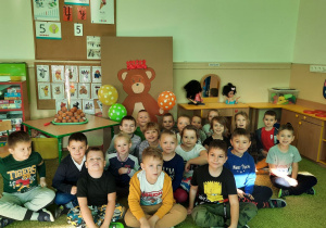 Dzieci siedzą przy misiu narysowanym na kartonie i świętują Światowy Dzień Pluszowego Misia.