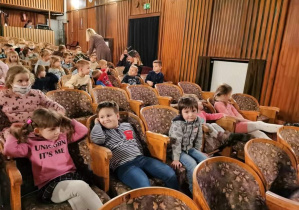 Dzieci siedzą na krzesłach w teatrze i czekają na spektakl.