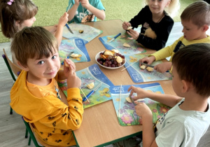 Dzieci siedzą przy stoliku i robią szaszłyki owocowe.