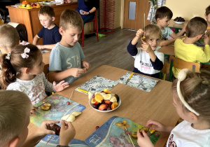 Dzieci siedzą przy stoliku i robią szaszłyki owocowe.