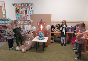 Dzieci stoją koło dużego narysowanego misia przed którym jest jego urodzinowy tort.