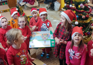 Dzieci w czerwonych strojach trzymają prezent od Mikołaja.