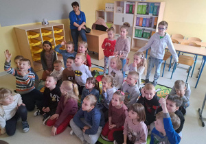 Dzieci siedzą na dywanie i odpowiadają na pytania nauczycielki szkoły.