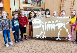 Dzieci stoją i trzymają w ręku duży szkielet dinozaura.