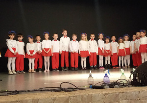 Dzieci ubrane w bało czerwone stroje śpiewają piosenkę o świętach wielkanocnych wraz z wielkanocnym zającem.