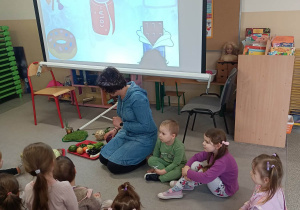 Dzieci siedzą na dywanie i oglądają prezentację na tablicy multimedialnej.
