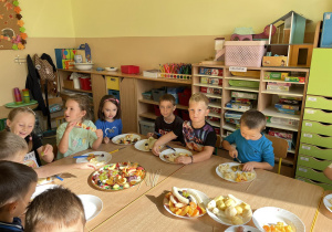 Dzieci siedzą przy stole i robią owocowe szaszłyki.